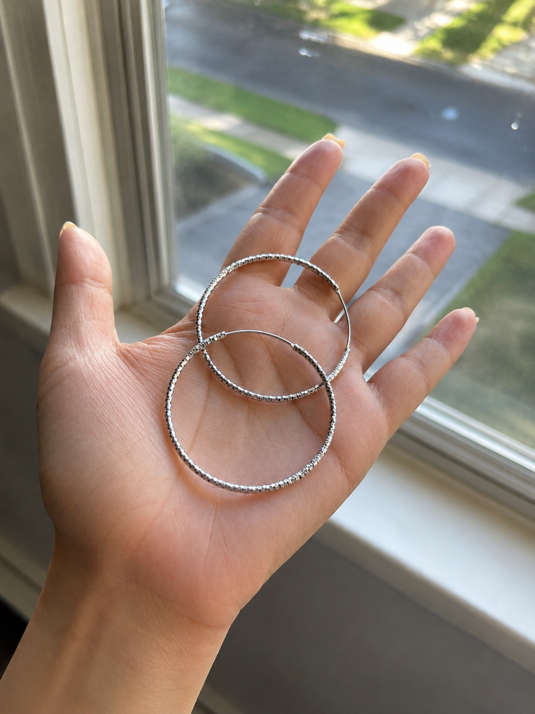 Hand holding two silver hoop earrings in front of a window