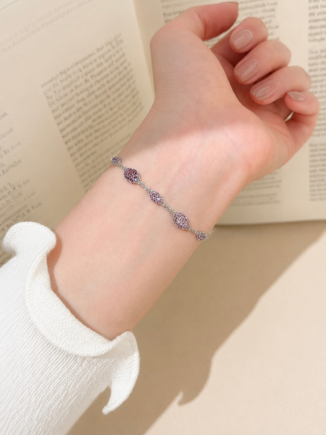 A delicate silver-toned chain bracelet with three oval purple gemstone charms, shown worn on a person's wrist against an open book background.