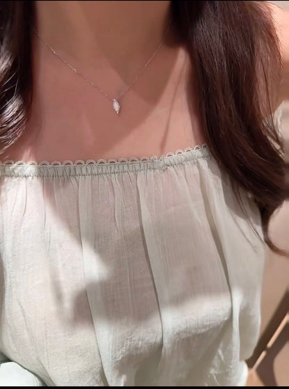 A close-up of a person wearing a delicate silver necklace with a small teardrop-shaped pendant, paired with a light beige off-the-shoulder top.