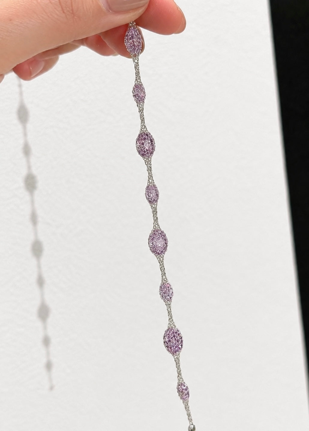A close-up of a hand holding a delicate silver-toned chain bracelet with multiple oval and round lavender-purple gemstones set in small woven bracelet, against a plain white background.