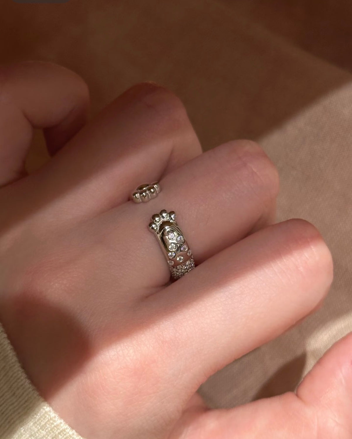 A silver ring with a small central charm and rows of clear stones, worn on a person's finger against a warm beige background.