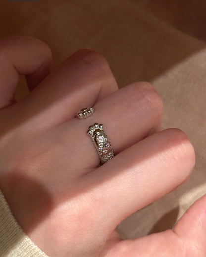 A silver ring with a small central charm and rows of clear stones, worn on a person's finger against a warm beige background.