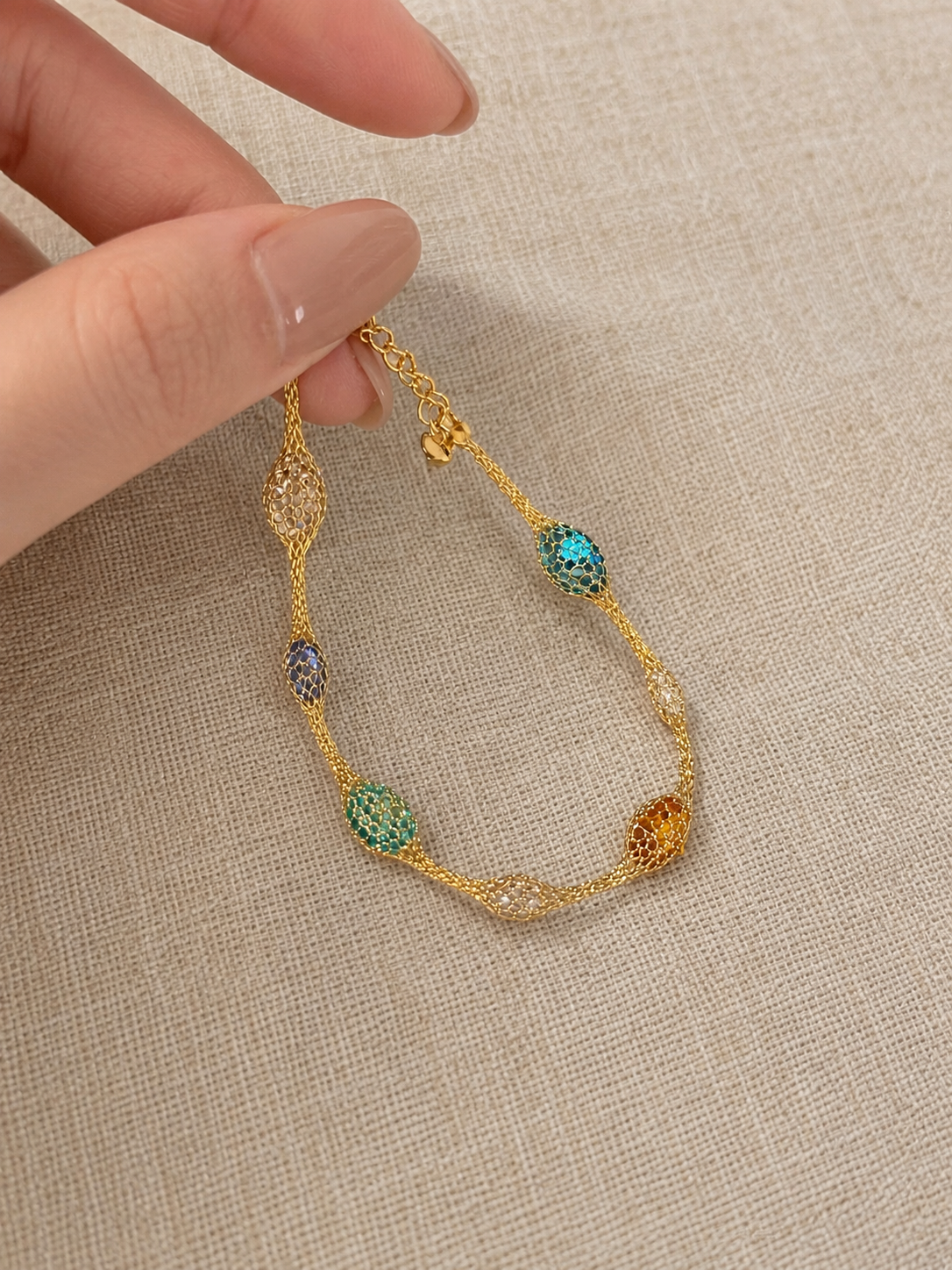 A gold chain bracelet with multiple textured oval links in turquoise, green, purple, and amber, held by a hand against a beige linen background.