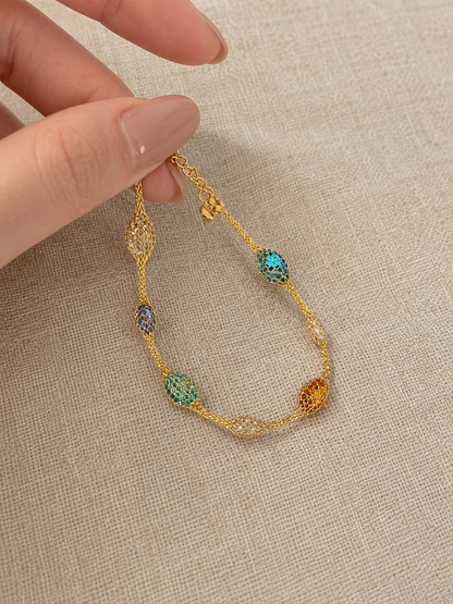 A gold chain bracelet with multiple textured oval links in turquoise, green, purple, and amber, held by a hand against a beige linen background.
