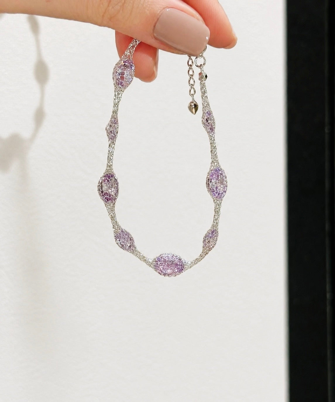 A silver-toned chain necklace with oval and marquise-cut light purple gemstones, held by a hand with beige manicured nails against a white textured background.