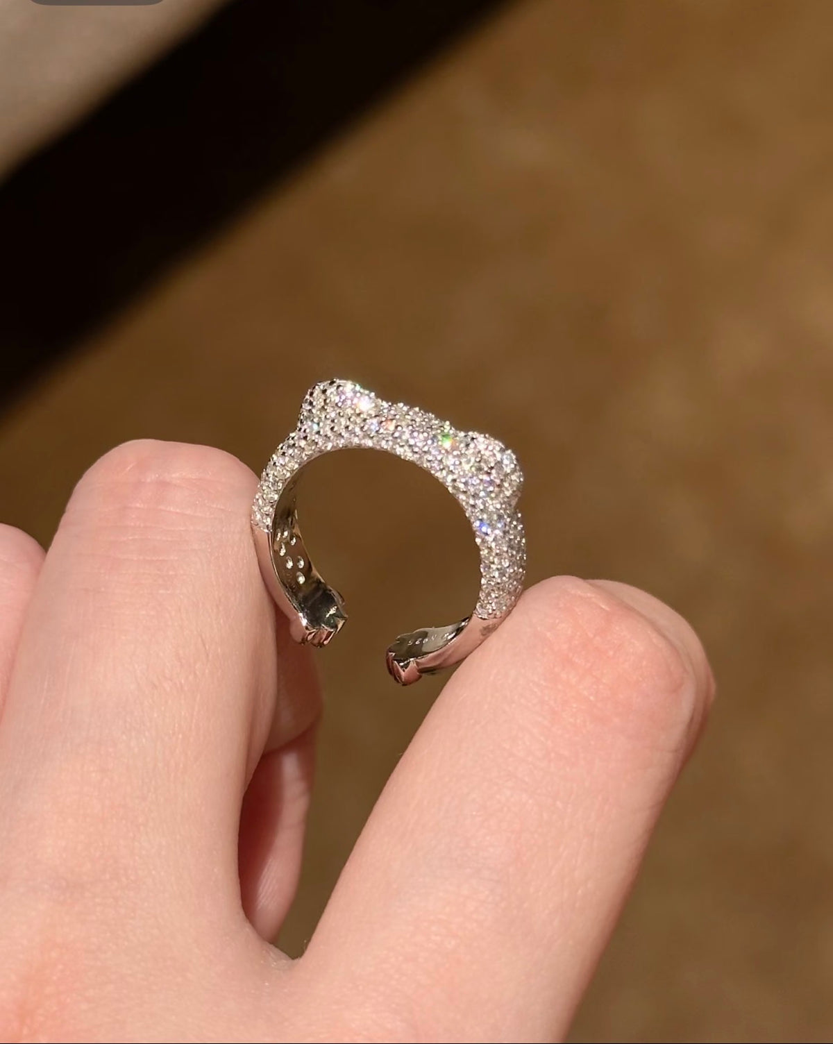 A silver-toned open ring adorned with pavé-set clear stones, held between fingers against a warm brown background.