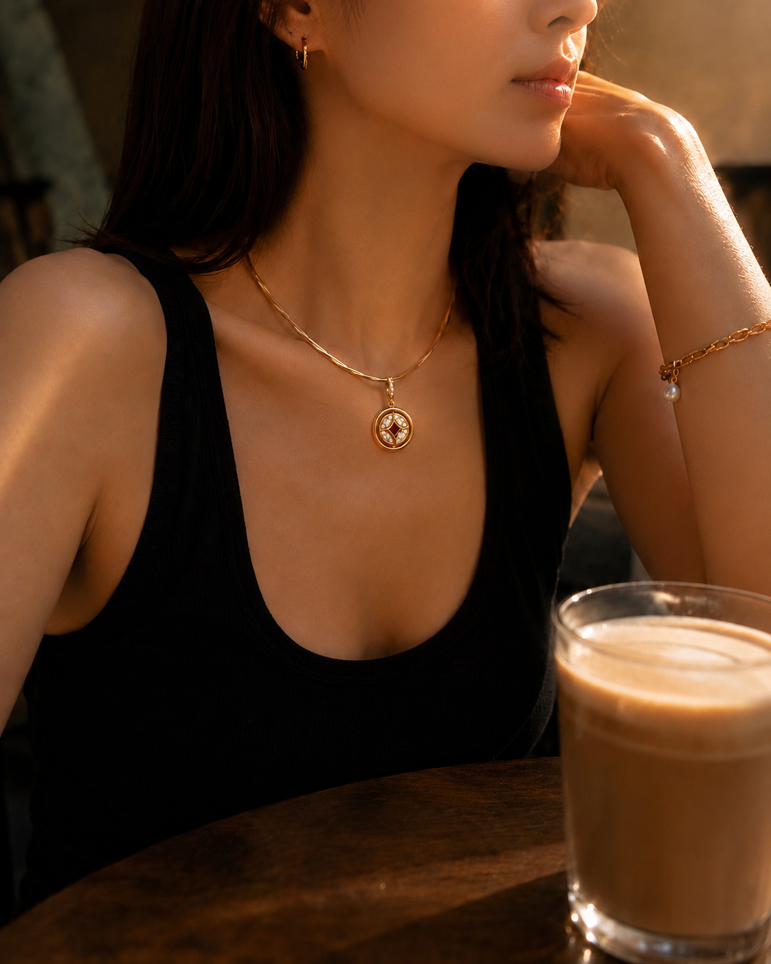 Woman in a black top with a gold necklace, sitting at a table with a glass of coffee.