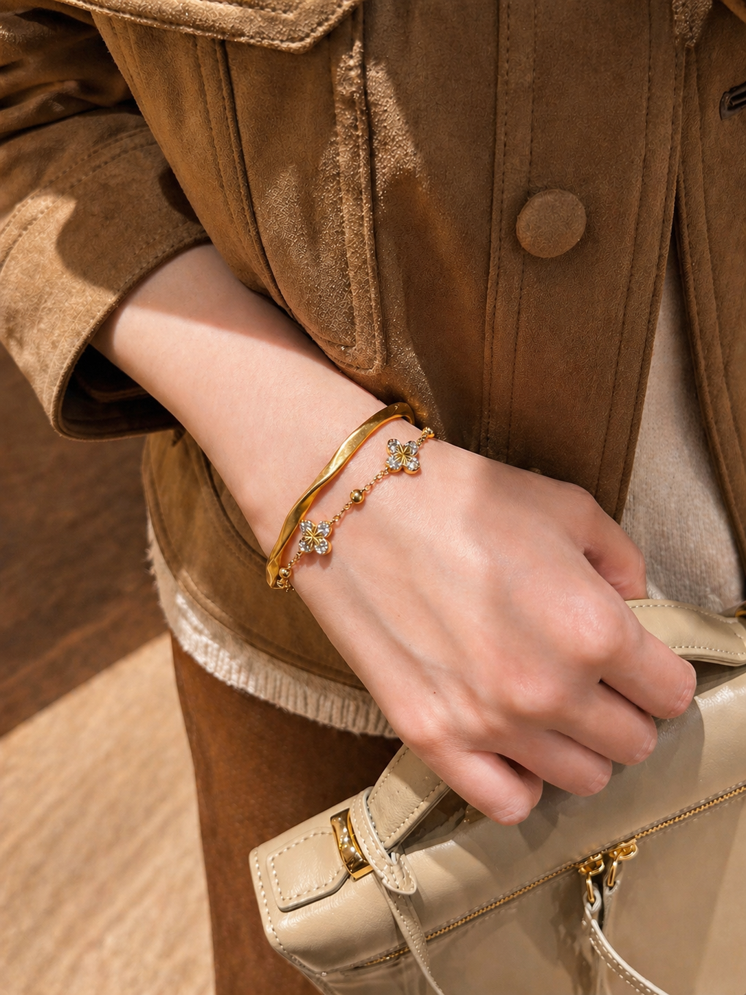 Hand wearing a gold bracelet with a beige handbag and brown coat