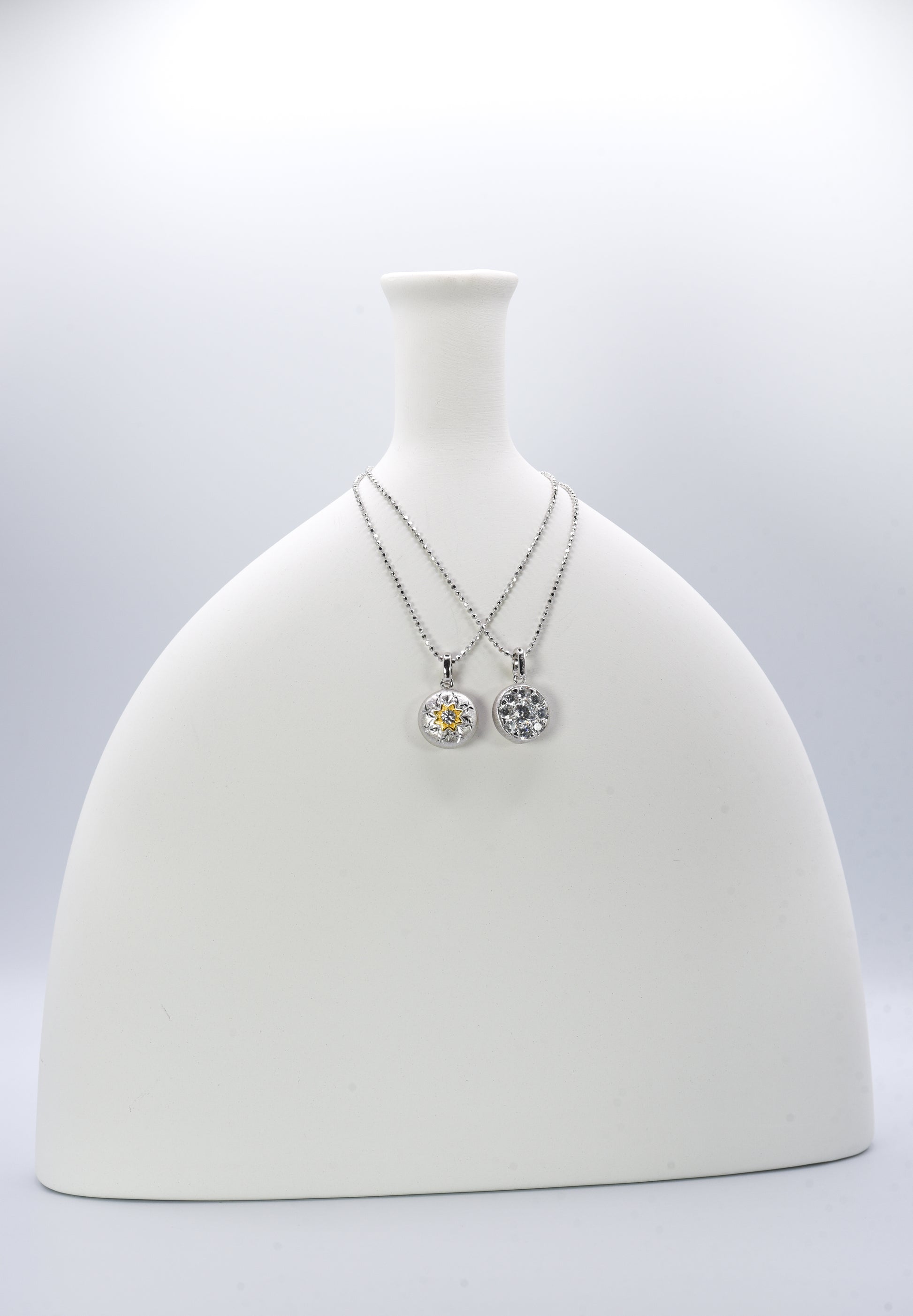 Two silver necklaces with round pendants on a white mannequin bust against a light gray background