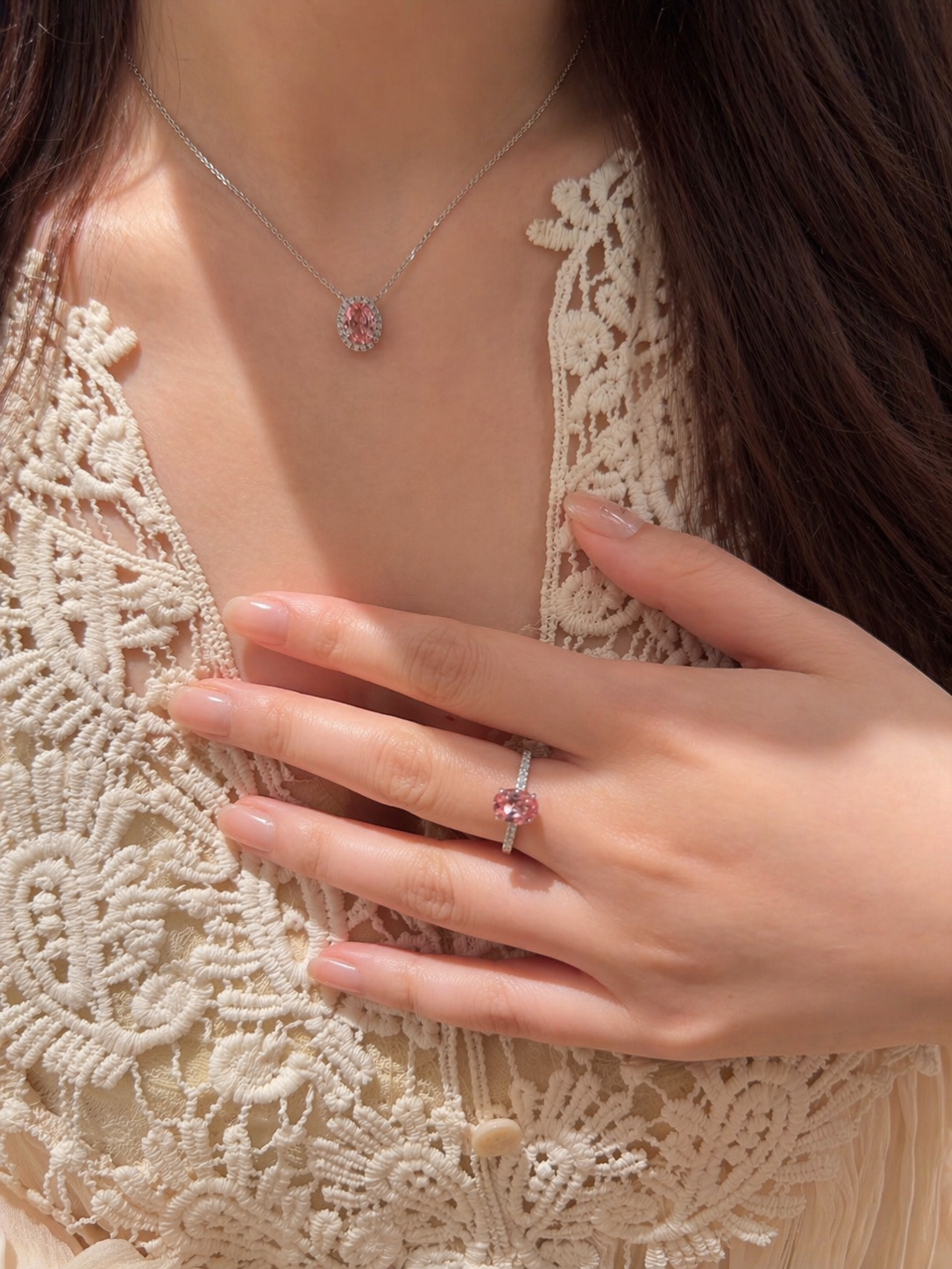 Close-up of a person wearing a silver necklace and ring with a pink gemstone on a beige lace garment.