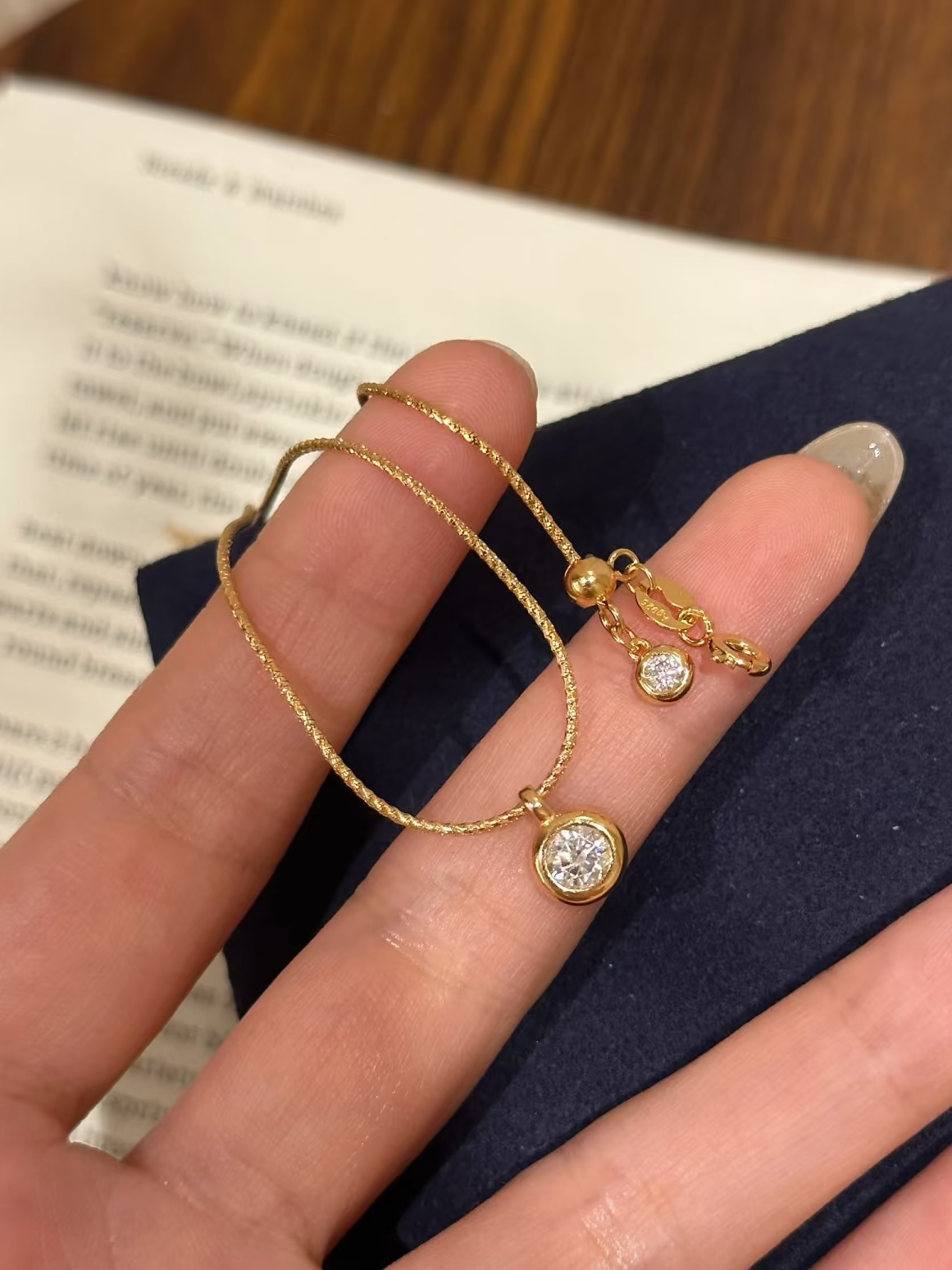 Gold bracelet with a diamond charm on a hand, against a book background