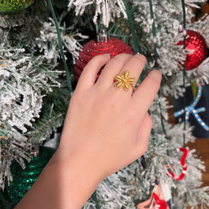Hand wearing a gold ring with a flower design, holding a red ornament against a Christmas tree background.