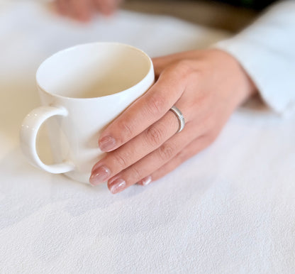 Hand holding a white mug on a light surface with a silver ring