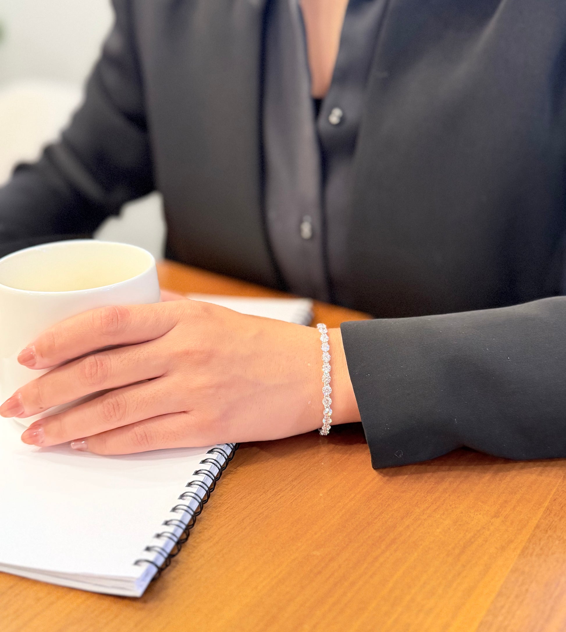 Person sitting at a desk with a notebook and a cup, wearing a bracelet.